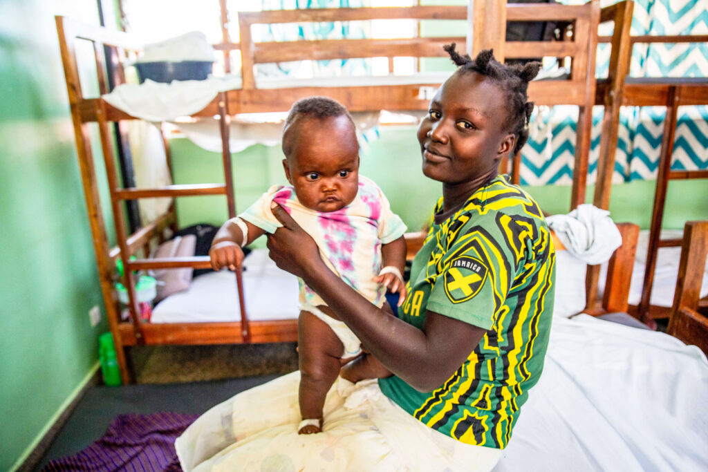 A woman with hair tied up, wearing a yellow and green shirt, sitting on a bed and holding a baby, in a hospital ward with wooden bunk beds in the background.