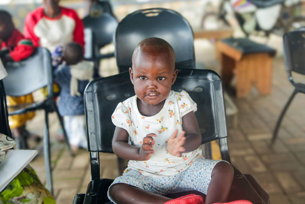 A smiling toddler wearing a floral top and patterned shorts sits on a black chair, clapping her hands.