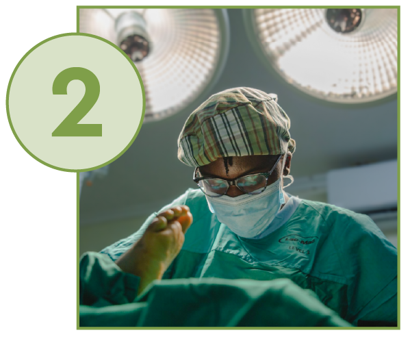  A medium-shot photograph taken in an operating room, showing a surgeon performing a procedure on a patient's foot. The surgeon is wearing a green surgical gown, a plaid scrub cap, a surgical mask, and glasses. They are intently focused on the patient's foot, which is elevated and prominent in the frame. Bright surgical lights are visible overhead, reflecting off the ceiling. A large, light-green circle with the number "2" inside overlays the top-left corner of the image.