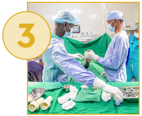 A wide-shot photograph taken in an operating room, showing two surgeons in light blue surgical gowns, caps, and masks working over a patient who is covered in green surgical drapes. The surgeon in the foreground, wearing long gloves, is reaching toward a tray of surgical instruments and supplies on a draped table in the bottom right. The second surgeon is standing directly opposite, focused on the patient's exposed limb (possibly an arm or leg) in the center. An anesthesiology monitor is visible in the background. A large, yellow-orange circle with the number "3" inside overlays the top-left corner of the image.