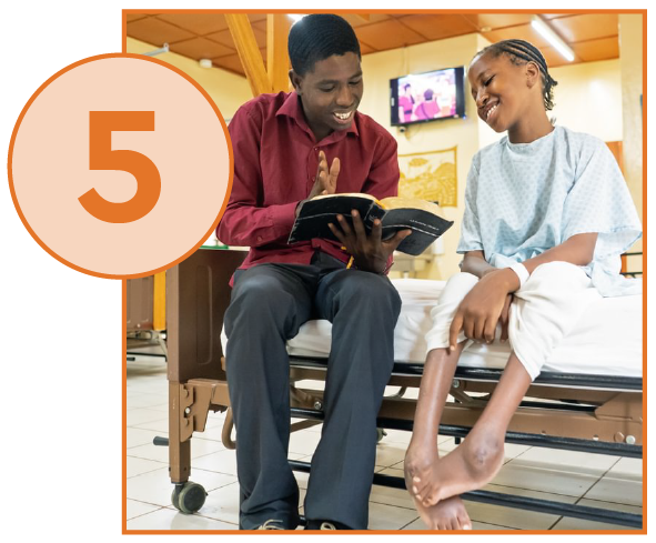  A photograph showing a young man sitting on the edge of a hospital bed next to a young female patient. The man is wearing a red collared shirt and black trousers, holding an open book, and smiling while looking at the girl. The girl is wearing a light blue hospital gown and is also smiling while looking at the man. Her bare legs and feet are visible, showing a healed or partially healed wound/scar near her ankle. The setting is a hospital ward with other beds and medical equipment visible in the background, and a TV screen mounted on the wall. A large, orange circle with the number "5" inside overlays the top-left corner of the image
